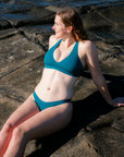 JAYD Swimwear model reclining against rocks on the beach, wearing the Vienna bikini top in cove green and matching Amalfi bottoms.