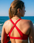 JAYD model viewed from behind wearing the Ruby Vienna bikini top with crisscross back straps, standing at the beach with the ocean and blue sky in the background.