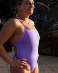 Model Kim wearing the purple lilac haze Zambia one piece swimsuit standing on a beach with large rocks in the background