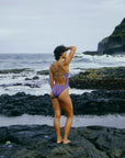 JAYD Swimwear model wearing the Zambia one piece in murasaki with contrasting olive shoulder straps and hip detailing, standing on rocky coastal terrain with ocean and cliffs in the background.