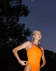 JAYD athlete Keira wearing the neon orange Zambia swimsuit standing outdoors at night with trees and a moonlit sky.