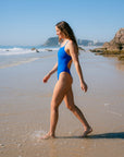 JAYD Swimwear model wearing the cobalt blue Zimmy one-piece swimsuit walking along the shallow surf of a sandy beach with ocean waves, rocks, and distant buildings in the background.