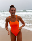 JAYD Swimwear features the Neon Orange Zimmy one piece from the front. This athletic one piece is a cheeky cut, it is being worn for leisure at the beach
