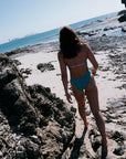 Person in an Atlantic coloured fixed back one piece standing on a rocky beach with ocean and skyline in the background. Airlie one piece offers contrasting strap detailing