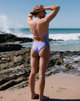 Woman in a Lilac Haze one piece standing on a rocky beach with ocean waves. The airlie one piece is built for performance