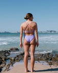 Woman in a lilac haze one piece standing on a rocky beach with ocean and city skyline in the background. She is wearing a full coverage one piece called Airlie