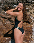 Woman in a black Lennox fixed back one piece with Moss green straps standing against a rocky background
