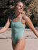 Woman wearing a matcha green Lennox one piece on a beach with rocks in the background