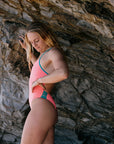 Woman in a vibe pink coloured one piece leaning against a stone wall. She is wearing the Lennox style, which is perfect for swimming