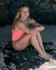 Woman in a JAYD Lennox one piece sitting on a rocky beach. She is wearing the colour vibe with atlantic hip cutouts and strap detailing