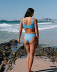 Woman in a JAYD blue bikini standing on a beach with ocean waves in the background. She is wearing the colour "Breeze"