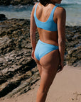 Person wearing a JAYD Breeze blue bikini standing on a rocky beach. She is wearing the Nalu top and the Tahiti bottoms