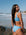 Carla wears JAYD Breeze blue bikini standing on a beach with ocean waves in the background. She is wearing the nalu top and tahiti bottoms