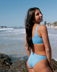 Carla wears JAYD Breeze blue bikini standing on a beach with ocean waves in the background. She is wearing the nalu top and tahiti bottoms