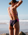 Woman in an Acai coloured IBIZA One Piece with Orchid cutouts, standing on a beach