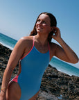 Woman in a JAYD breeze blue and vibe pink one piece called IBIZA standing on a rocky beach with clear blue sky.