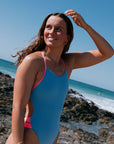 Woman in a breeze blue IBIZA one piece standing on a rocky beach with clear blue sky.