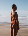 Charlotte in a Cinnamon (rust-brown) swimsuit standing on a beach with ocean waves in the background