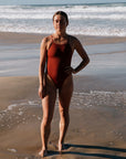 Woman in a Tallow swimsuit by JAYD Swimwear, standing on a beach with ocean waves in the background