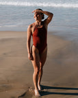 Charlotte in a TALLOW swimsuit in Cinnamon, standing on a beach with ocean waves in the background