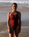 Charlotte wears the Tallow One Piece by JAYD Swimwear in shade Cinnamon, standing on a beach with ocean waves in the background