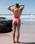 Woman in a JAYD Tallow one piece standing on a beach with ocean waves in the background. She is wearing the colour Vibe.