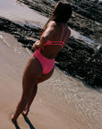 Woman in a vibe coloured one piece standing on a beach with water and rocks in the background. Vibe is a bright pink coloured