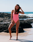 Woman in a pink, vibe coloured one piece by JAYD. Standing on a rocky beach with ocean waves in the background. She is ready to dive in with confidence