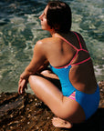 Woman in a cancun blue swimsuit sitting on a rock by the water. She is wearing the Zambia one piece and is ready to dive in with confidence