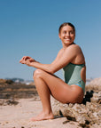 Woman in a matcha green fixed back on piece sitting on a rock at the beach. She is wearing the Zambia style