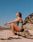 Woman in a matcha green fixed back one piece sitting on a rock at the beach with clear blue sky.