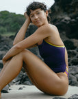 JAYD model Sophie sitting on a rocky beach wearing the Lennox one piece in Acai with Daffodil yellow straps and multi-coloured hip cut-outs.