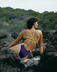 JAYD Swimwear model sitting on a rocky beach in the Lennex contrast one piece in Acai purple and daffodil yellow.