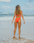 JAYD swimwear model standing on the beach facing the water in the Airlie Blaze one piece swimsuit.