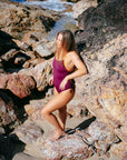 Model standing on a rocky beach wearing the full coverage, high performance JAYD Swimwear Airlie one piece in sangria with contrasting flare shoulder straps.