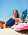 Athlete Liv Corrin in the navy blue Albi one piece swimsuit sitting on a beach leading on her pink board with tall buildings in the background.