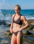 JAYD athlete Dom Stitt modeling the Cacao Amalfi bottoms on a rocky beach with ocean waves and water splashes in the background.