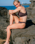 JAYD model Dom sitting on dark rocks by the ocean, wearing the Cacao Amalfi bikini bottoms. The background features blue water and sky with a distant lighthouse.