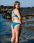 JAYD Swimwear model stands in shallow water at a rocky beach wearing the Amalfi bikini bottoms in cove, photographed from the back and side.