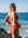 JAYD Swimwear model Jemima wearing the Amalfi bikini bottoms in ruby red, stands on a rocky beach with the ocean and blue sky behind her, looking over her shoulder.
