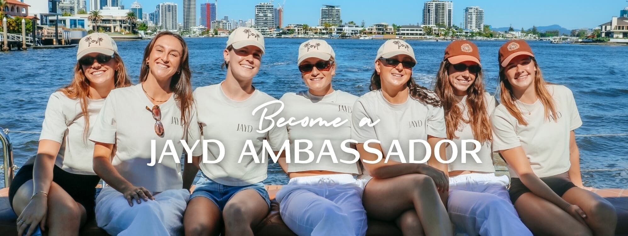 Seven JAYD Swimwear athletes & ambassadors sitting together on a boat or docked vessel with water and a city skyline behind them. Large overlaid text reads 'Become a JAYD AMBASSADOR.