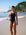 JAYD Swimwear athlete Claudia Slaven wearing the black Apsley one-piece swimsuit with bright pink sorbet and rave green shoulder straps walks on a sandy beach with ocean waves and trees in the background.