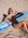 JAYD Swimwear model Cyra wearing the Apsley Cacao swimsuit with white shoulder straps, reclines on a blue and white ski on a sandy beach, shielding her eyes from the sun.