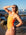 JAYD Swimwear surf life saving athlete Mia Cook standing on a rocky beach shading her eyes from the afternoon sun, wearing the Apsley one piece swimsuit in neon orange.