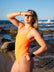 JAYD Swimwear surf life saving athlete Mia Cook standing on a rocky beach shading her eyes from the afternoon sun, wearing the Apsley one piece swimsuit in neon orange.