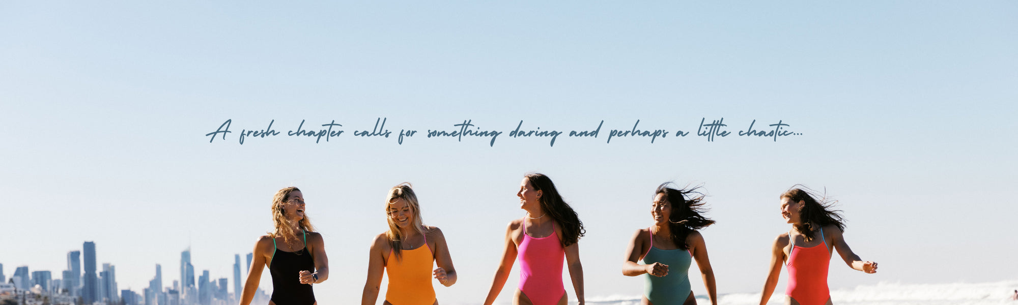 Five women in colorful swimsuits walking on a beach with a city skyline in the background.