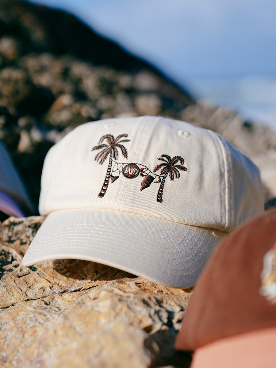 Cream JAYD Swimwear baseball hat with brown palm trees & swimsuit embroidery sitting on a rock.