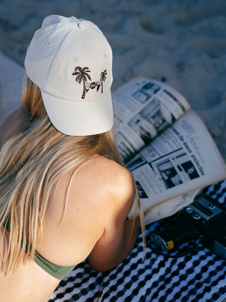 JAYD model laying on the beach reading a magazine wearing the cream JAYD baseball cap.