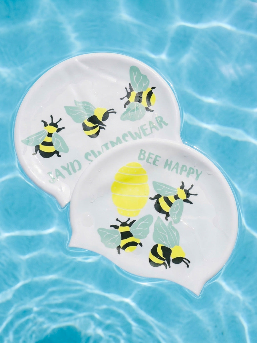 Two white swim caps with yellow and black bumblebee illustrations and the words 'JAYD SWIMWEAR' and 'BEE HAPPY', floating on blue water.