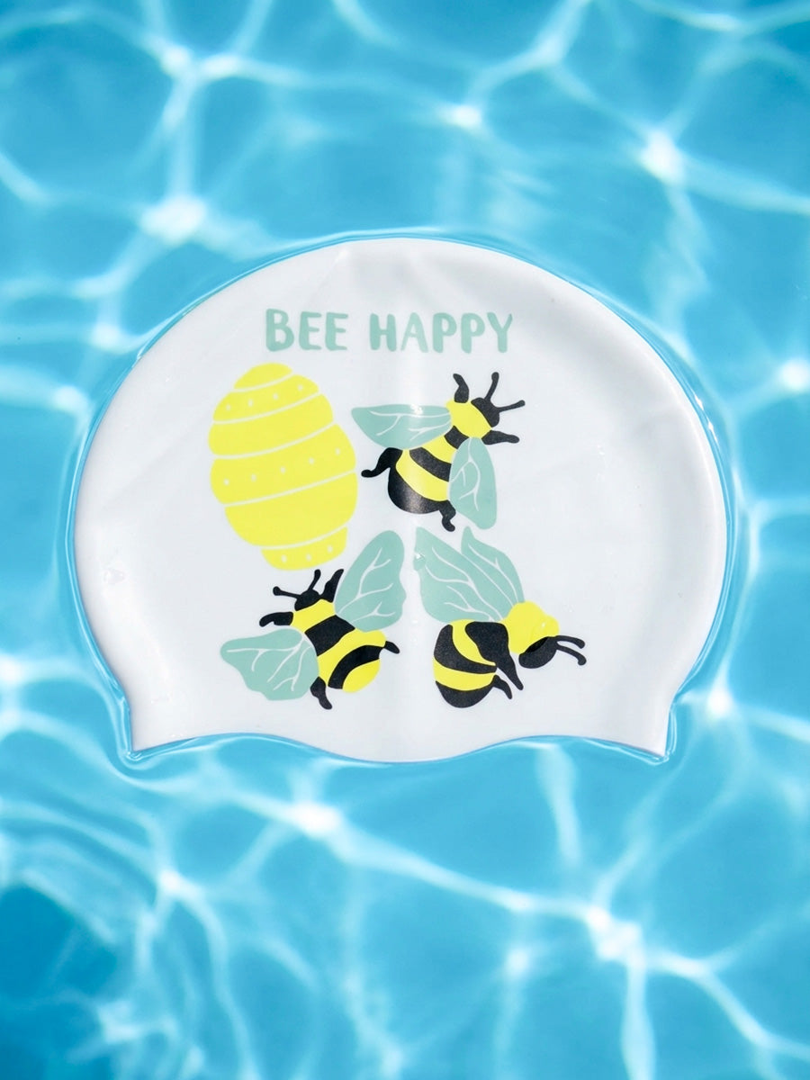 A white swim cap featuring a cute 'BEE HAPPY' design with yellow bees, a yellow honeycomb, and green leaves, shown floating on a bright blue water background with ripples.